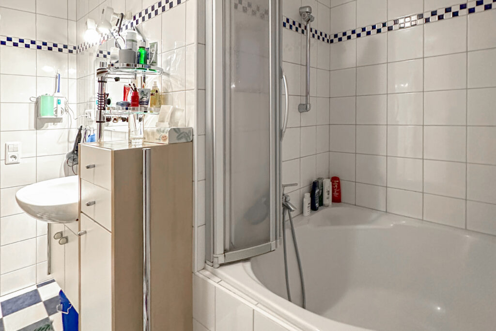 Vorher: Bad mit weißen Fliesen und blauer Bordüre, Eckbadewanne mit Duschabtrennung, Waschbecken und hohem Badschrank.