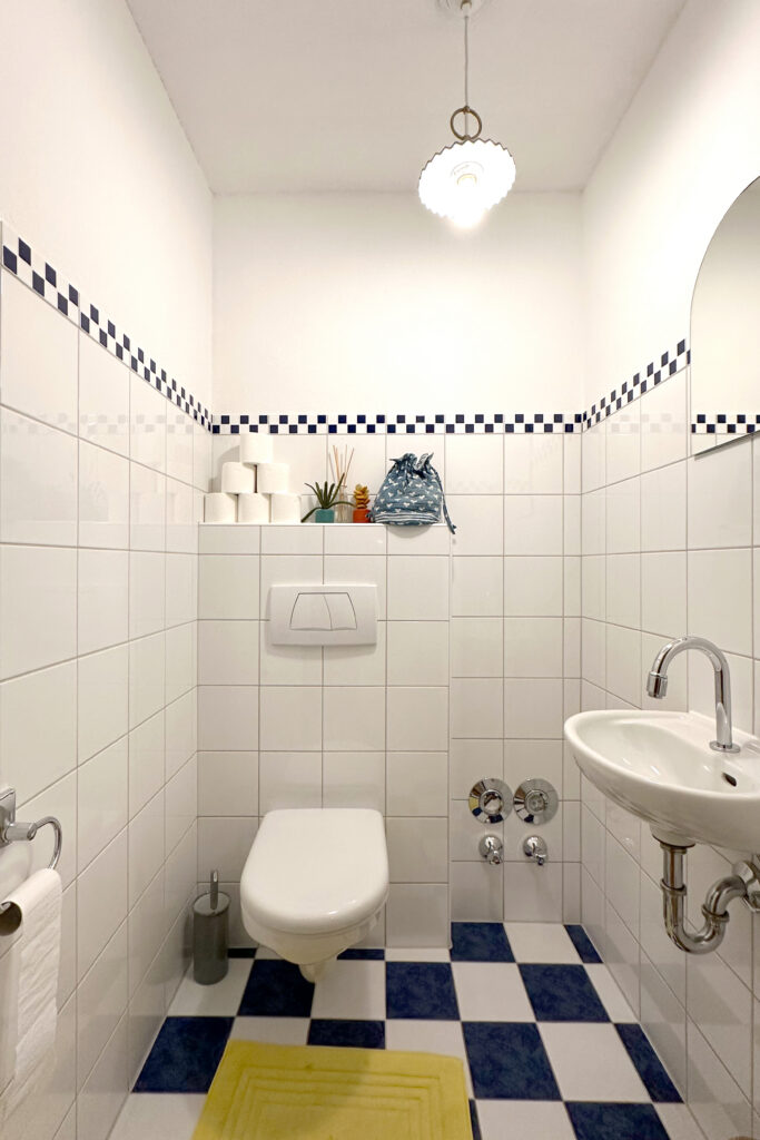 Vorher: schmales WC mit weißen Fliesen, blauer Mosaikbordüre, blau-weiß kariertem Boden und kleinem Handwaschbecken.